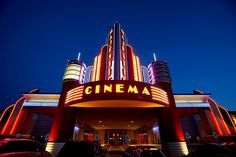 the cinema sign is lit up in red and blue lights at night time, with cars parked below it