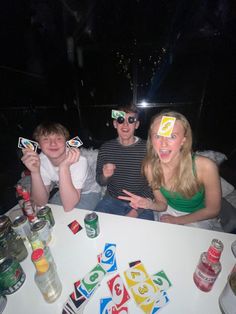 three people sitting at a table with beer glasses on their heads and playing cards in front of them