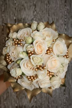 a bridal bouquet with white roses and gold accents on a wooden table next to a person's hand