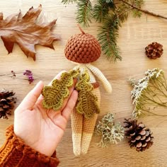 a hand holding a knitted doll next to pine cones and leaves