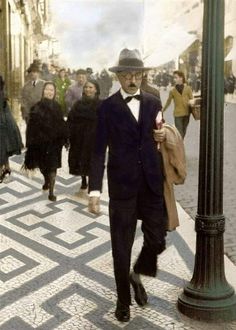 an old man in a suit and hat walking down the street with people behind him