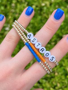 "Cheer on your favorite baseball team with this Astros gold bead bracelet set! Message me if you're interested in a set for a different team! Details: -Set includes three bracelets: Astros letter bracelet, orange color strip bracelet, blue color strip bracelet -14k gold filled beads will not tarnish -6.5\" long -3mm gold beads -7mm alphabet beads" Gold Heishi Beads Friendship Bracelet, Gold Stretch Bangle With Letter Beads, Gold Stretch Bangle Bracelet With Letter Beads, Gold Heishi Beads Bracelets For Friendship, Gold Heishi Beads Bracelet Gift, Gold Heishi Beads Bracelets As Gift, Personalized Heishi Beads Jewelry For Friendship, Friendship Jewelry With Letter And Heishi Beads, Customizable Heishi Bead Bracelets