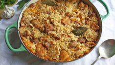 a green pot filled with rice and meat on top of a white table cloth next to silver spoons