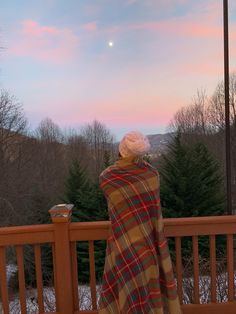 a woman wrapped in a blanket looking out over the mountains at sunset on a deck