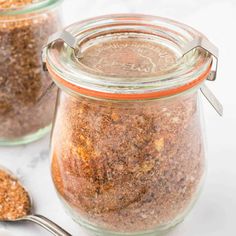 two jars filled with spices next to spoons on a white counter top and one jar has seasoning in it