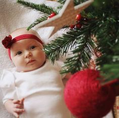 a baby laying under a christmas tree next to a red ornament and ornaments