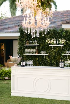 a white bar with wine glasses and bottles on it in front of a house that has a chandelier hanging from the ceiling