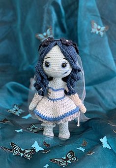 a crocheted doll is standing on a blue sheet with butterflies around it and wearing a dress