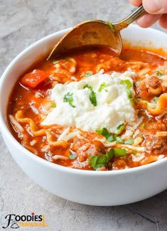 a bowl of pasta soup is being spooned with a ladle and topped with sour cream