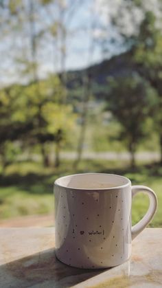 a white coffee cup sitting on top of a table next to a forest filled with trees