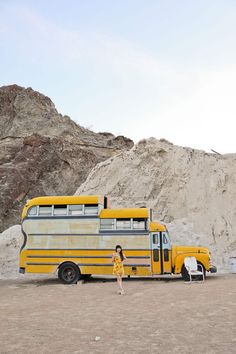 a woman standing in front of a yellow bus on the side of a mountain range