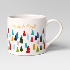 a white coffee mug with christmas trees on the front and merry and bright written in gold