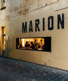 people are sitting in the window of a store called marion, on a brick sidewalk