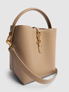 Find SAINT LAURENT Le 37 Bucket Bag on Editorialist. The SAINT LAURENT Le 37 Bucket Bag features a top handle and an adjustable, detachable strap. The bucket bag is made of leather and has gold-toned hardware. It measures 20cm in height, 17cm in width, and 13cm in depth. The strap drop is 45cm and the handle drop is 6cm. The bag has a top strap with a metal logo hook closure. It is a small bucket bag that can be hand carried, worn on the shoulder or worn crossbody. Small Bucket Bag, Beige Bag, Mini Bucket Bags, Ski Accessories, Bags Logo, Dark Beige, Leather Bucket Bag, Leather Bucket, Metal Logo