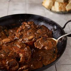 a skillet filled with meat and mushrooms on top of a tile floor next to bread