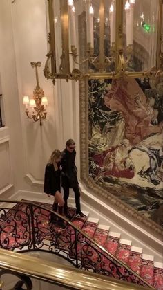 two people walking up the stairs in an ornate room with chandelier and tapestry