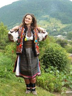 a woman standing on top of a lush green hillside wearing a colorful dress and jacket