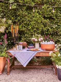 a table with flowers and candles on it in front of a bushy wall, surrounded by potted plants