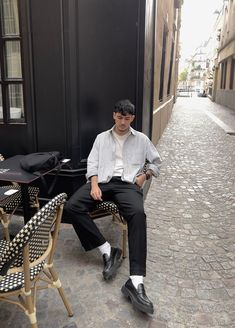 Boy sitting on a Parisian street chair in Le Marais, wearing a striped button-down shirt, pleated trousers from Uniqlo, and black shoes, against a stone street backdrop. Men Loafers Outfit, Black Trousers Outfit, Loafer Outfits, Loafers Men Outfit, Outfits With Striped Shirts, Black Loafers Men, Black Trousers Men, Parisian Street, Minimalist Fashion Men