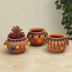 three clay pots with colorful beads and tassels sit on a table next to a potted plant