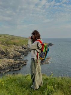 a woman standing on top of a lush green hillside next to the ocean holding a camera