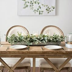 a wooden table topped with white plates and candles next to a plant filled centerpiece