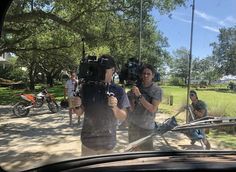 two men are filming in the street while others look on from behind a car's side view mirror