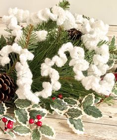 white cotton flowers and pine cones are arranged on a wooden table with holly berries, evergreen leaves and pine cones