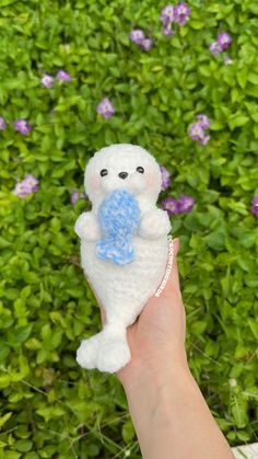 a hand holding a small white teddy bear in front of some purple and green plants