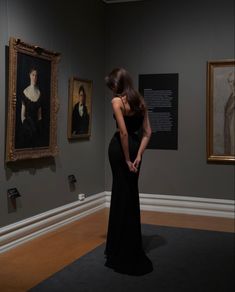 a woman in a black dress looking at some paintings on the wall and holding her hands behind her back