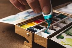 a person holding a paintbrush in their hand near an open box of watercolors