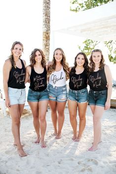 four girls in black shirts and denim shorts standing on the beach with palm trees behind them