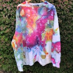 a colorful tie dye sweatshirt hanging on a wooden hanger in front of some bushes