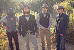 three men wearing hats and vests standing on a dirt road in front of bushes