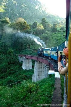 a blue train traveling over a bridge next to lush green mountains