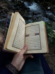 a person is holding an open book in their hand while sitting on the ground next to a stream