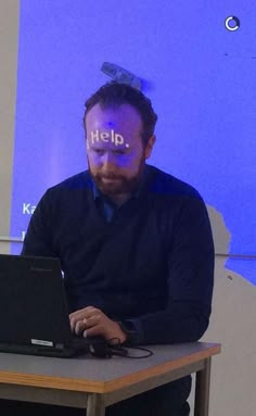 a man sitting in front of a laptop computer on top of a table with the word help projected on his face