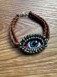 Handmade Evil Eye bracelet, featuring a turquoise embroidered eye surrounded by glass beads and rhinestones. Medium Evil Eye bracelet with braided leather, clasp and leather back. Overall length is 7". Eye is approx. 1.88" L x .75" W. The evil eye is a curse thought to be given by a malicious glare that can cause bad luck or loss. Wearing the evil eye wards of the curse protecting you throughout your day.   Necklace handmade by a high school art teacher and a woman owned small business. Details: Bohemian Handmade Evil Eye Bracelet For Festivals, Handmade Adjustable Amulet Beaded Bracelets, Beaded Bracelet Jewelry For Good Luck, Good Luck Beaded Bracelet Jewelry, Handmade Turquoise Bohemian Evil Eye Bracelet, Bohemian Beaded Turquoise Evil Eye Bracelet, Good Luck Beaded Bracelet, Bohemian Jewelry With Rhinestones For Festivals, Bohemian Festival Jewelry With Rhinestones