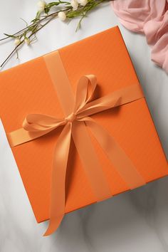 an orange gift box with a bow on it next to some flowers and a pink rose