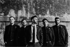 a group of men standing next to each other in front of a brick wall wearing suits and ties