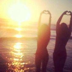 two people standing in the water making a heart shape with their hands while the sun is setting