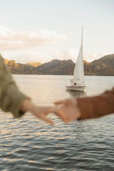 two people holding out their hands in front of a sailboat on the water with mountains in the background