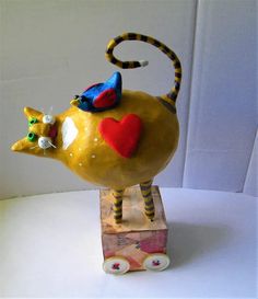 a yellow teapot with a red heart on it's head and legs, sitting on top of a wooden block