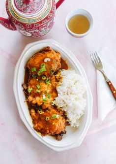 a white plate topped with meat and rice next to a tea pot on top of a table