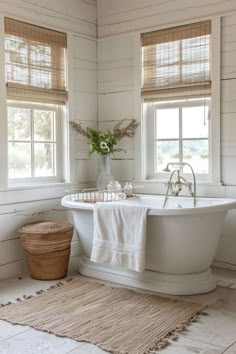 a white bath tub sitting in a bathroom next to two windows and a rug on the floor