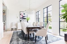 a dining room table with white chairs in front of large windows and an area rug on the floor