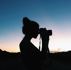 a woman holding a camera up to her face in front of the sun at dusk