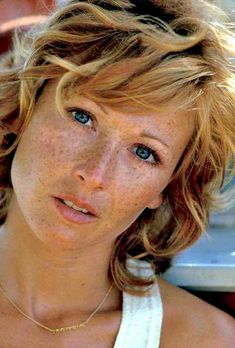 a woman with freckled hair and blue eyes looks at the camera while wearing a white halter top