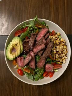 a white bowl filled with steak, corn and salad on top of a wooden table