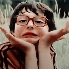 a young boy wearing glasses sitting in front of tall grass with his hands on his face